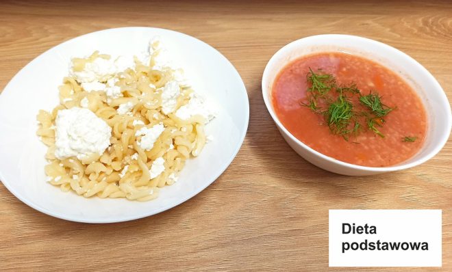 Na zdjęciu przedstawiono posiłek oznaczony jako „Dieta podstawowa”. Na pierwszym talerzu znajdują się makaron świderki z dodatkiem białego twarogu. Obok stoi miseczka z zupą pomidorową ozdobioną świeżym koperkiem. Całość ustawiono na jasnym drewnianym blacie