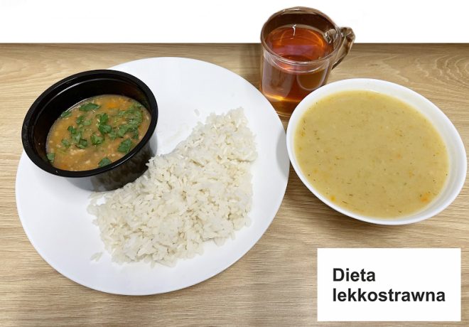 Zdjęcie ukazuje zestaw obiadowy oznaczony jako „dieta lekkostrawna”. Na talerzu podano porcję białego ryżu oraz mały pojemnik z sosem warzywno-mięsnym posypanym świeżą zieleniną. Obok znajduje się miska z zupą kremem o jasnej barwie oraz przezroczysta szklanka z herbatą