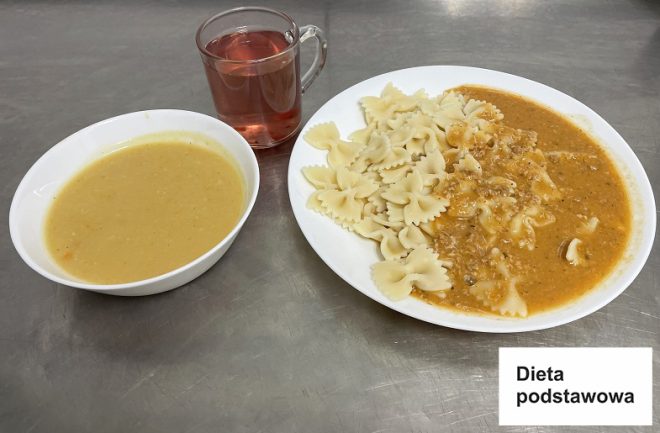 Na metalowym blacie znajduje się zestaw obiadowy w ramach diety podstawowej, co wskazuje widoczna w rogu kartka z napisem: „Dieta podstawowa”. Na dużym talerzu podano makaron kokardki z sosem mięsnym. Obok znajduje się miseczka z zupą kremem z warzyw, a w przezroczystym kubku nalano napój w kolorze różowym