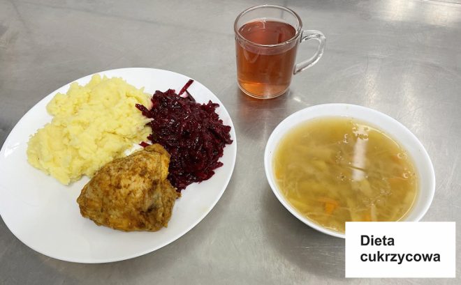 Na zdjęciu widoczne jest danie obiadowe przygotowane w ramach diety cukrzycowej. Na białym talerzu znajdują się ziemniaki puree, buraczki na ciepło oraz porcja pieczonego mięsa. Obok talerza stoi przezroczysta szklanka z herbatą, a w oddzielnej misce zupa z kawałkami warzyw na jasnym wywarze. W prawym dolnym rogu zdjęcia znajduje się biała kartka z napisem „Dieta cukrzycowa”. Całość podana jest na metalowym blacie