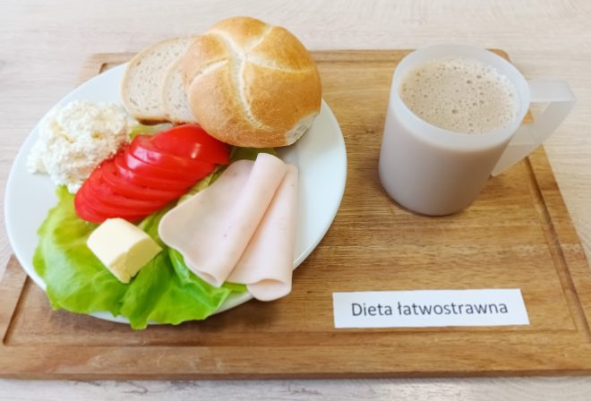 Na drewnianej podstawce umieszczono zestaw śniadaniowy oznaczony jako "Dieta łatwostrawna". Na talerzu leżą plasterki pomidora, szynki drobiowej, porcja twarogu, liść sałaty, kawałek masła oraz bułka pszenna. Obok talerza stoi biały kubek z kawą z mlekiem lub innym napojem mlecznym