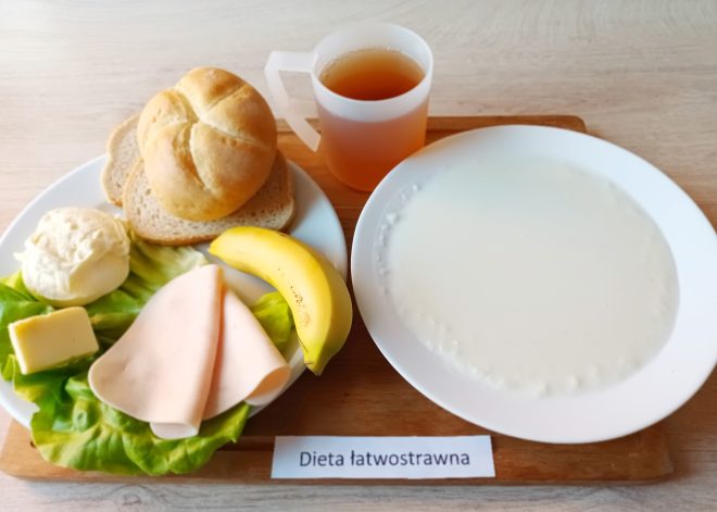 Zestaw śniadaniowy oznaczony jako "Dieta łatwostrawna", prezentowany na drewnianej podstawce. Na białym talerzu podano plaster szynki drobiowej, kawałek żółtego sera, porcję białego twarogu, sałatę oraz połowę banana. Obok znajdują się bułka kajzerka i dwie kromki chleba. Na drugim talerzu widoczna gładka, biała kasza manna. Całość uzupełnia przezroczysty kubek z ciemnym napojem, prawdopodobnie herbatą