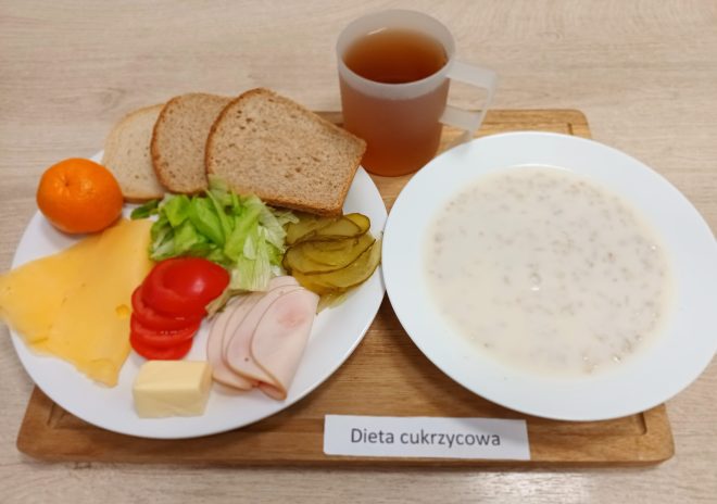 Zdjęcie przedstawia zestaw śniadaniowy oznaczony jako "Dieta cukrzycowa". Na drewnianej tacce znajduje się kilka elementów: talerz z różnorodnymi składnikami, miska z mleczną owsianką oraz kubek z herbatą. Na talerzu widoczne są trzy kromki jasnego pieczywa, plastry żółtego sera, sałata lodowa, plasterki pomidora, ogórki kiszone, zwinięte plasterki wędliny drobiowej oraz kostka masła. Obok znajduje się mandarynka jako porcja owocu. Wszystkie składniki ułożone są estetycznie i zachęcająco, podkreślając zróżnicowanie i zbilansowanie posiłku zgodnego z wymogami diety dla diabetyków