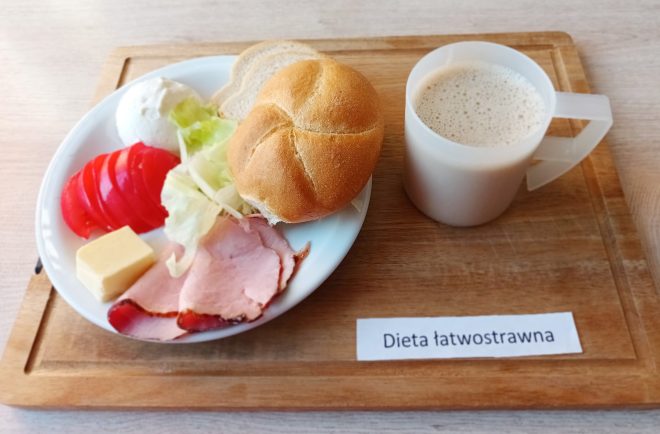 Zdjęcie przedstawia śniadanie z etykietą "Dieta łatwostrawna". Na białym talerzu znajduje się świeża bułka pszenna oraz dwa cienkie plasterki chleba. Obok położono plastry szynki, kilka kawałków pomidora, małą porcję sałaty oraz kostkę żółtego sera. Na talerzu widać także dwie porcje białego twarożku. Obok talerza stoi biały kubek z mlekiem lub napojem mlecznym. Wszystko ułożone jest na drewnianej desce z widoczną etykietą "Dieta łatwostrawna"