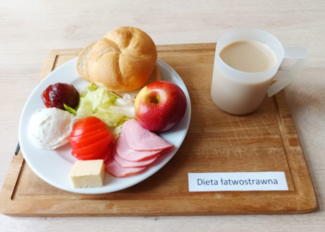 Na zdjęciu przedstawiono posiłek oznaczony jako „Dieta łatwostrawna”. Na białym talerzu znajdują się bułka kajzerka, plasterki czerwonego pomidora, liście sałaty, jabłko, dwa plasterki szynki, porcja twarogu, kostka żółtego sera oraz łyżeczka konfitury. Obok talerza stoi przezroczysty kubek wypełniony kawą z mlekiem. Całość ustawiono na drewnianej tacce, na której znajduje się napis „Dieta łatwostrawna”