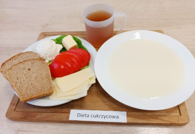 Na drewnianej tacy znajduje się śniadanie oznaczone jako „Dieta cukrzycowa”. Na talerzu po lewej stronie umieszczono dwa kawałki chleba pełnoziarnistego, biały serek, plaster masła, plasterki pomidora, sałatę oraz plasterki żółtego sera. Po prawej stronie znajduje się talerz z jasną, kremową zupą mleczną. Obok stoi przezroczysty kubek z herbatą