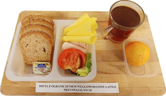 Zdjęcie przedstawia śniadanie opisane jako "dieta z ograniczeniem węglowodanów łatwo przyswajalnych". Zestaw zawiera plastikowy pojemnik z trzema kromkami pełnoziarnistego chleba oraz małym opakowaniem masła Lacima. Obok znajdują się składniki na kanapki: zwinięte plastry żółtego sera, dwa plastry drobiowej wędliny, kawałek pomidora oraz liść sałaty. Zestaw uzupełnia szklany kubek z herbatą oraz plastikowy pojemnik z pomarańczą. Na desce widnieje papierowa etykieta z pełnym opisem diety