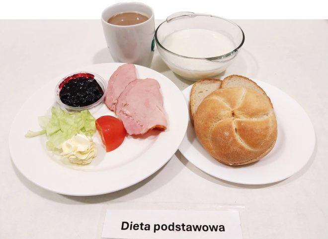 Na białym tle znajduje się zestaw śniadaniowy oznaczony jako "Dieta podstawowa". Na jednym talerzu podano plasterki wędliny, pomidora, sałatę, masło oraz niewielką porcję dżemu w plastikowym pojemniku. Obok znajduje się bułka pszenna i kromki chleba, a w tle widoczny jest kubek z kawą z mlekiem oraz szklana miseczka z jogurtem naturalnym