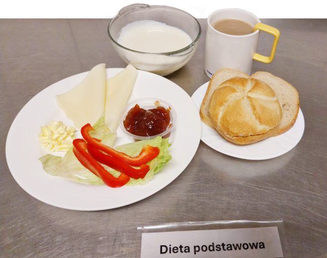 Na zdjęciu przedstawiono posiłek oznaczony jako „Dieta podstawowa”. Na białym talerzu ułożono dwa plastry sera żółtego, liście sałaty, paski czerwonej papryki oraz porcję konfitury w małym pojemniczku. Towarzyszy im bułka kajzerka z kromkami chleba oraz szklanka z kawą w żółtym kubku. W tle widoczna jest miseczka z mlekiem