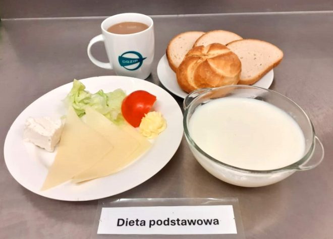 Zestaw śniadaniowy oznaczony jako "Dieta podstawowa". Na talerzu znajdują się dwa plastry żółtego sera, liść sałaty, plaster pomidora oraz porcja dżemu w małym pojemniku. Na talerzu obok ułożono bułkę kajzerkę i dwie kromki chleba. W tle przezroczysta miska z mlekiem oraz niebieski kubek z kawą