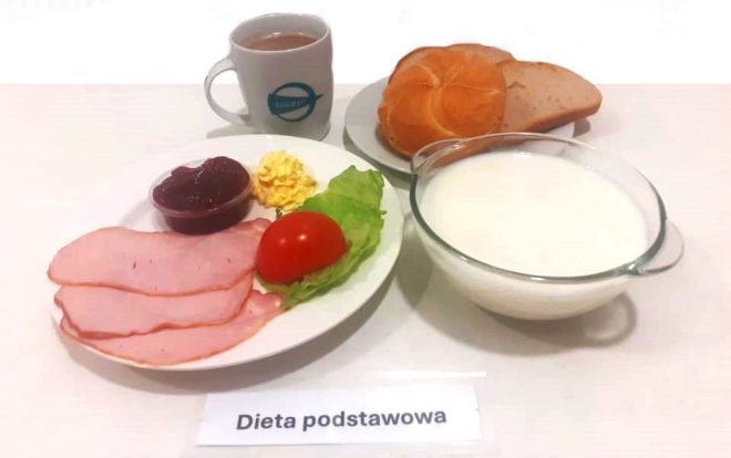 Na zdjęciu znajduje się zestaw śniadaniowy oznaczony jako "Dieta podstawowa". Na białym talerzu znajdują się trzy plastry szynki, liść sałaty, plaster pomidora, porcja pasty jajecznej oraz mały pojemniczek z konfiturą. Obok stoi filiżanka kawy z mlekiem, bułka kajzerka i kromka chleba. Na stole umieszczono także przezroczystą miskę z mlekiem