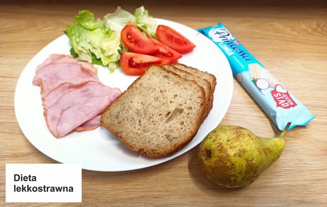 Zdjęcie przedstawia śniadanie oznaczone jako "Dieta lekkostrawna". Na białym talerzu ułożono trzy kromki jasnego chleba pełnoziarnistego, kilka plastrów szynki drobiowej, liście sałaty lodowej oraz pokrojone kawałki pomidora. Obok talerza leży gruszka oraz batonik w opakowaniu. Na dole zdjęcia znajduje się etykieta z napisem "Dieta lekkostrawna". Wszystko umieszczone jest na drewnianym blacie