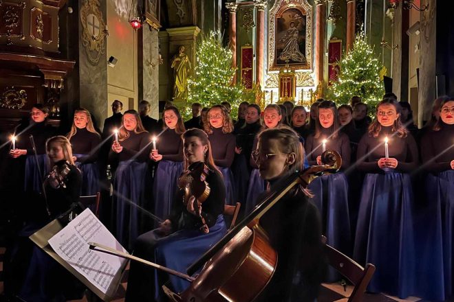 Młode artystki z chóru i orkiestry, trzymające zapalone świece, stojące na tle oświetlonego ołtarza i choinek. Na pierwszym planie widoczne są muzycy grający na wiolonczeli i skrzypcach, co podkreśla kameralną atmosferę koncertu