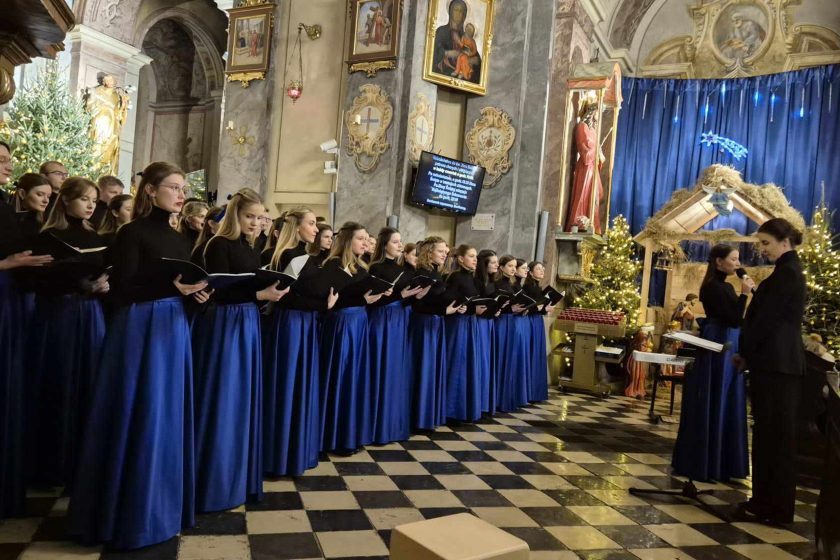 Ujęcie chóru w pełnym składzie, stojącego w eleganckich strojach – długich granatowych spódnicach i czarnych bluzkach. W tle widać szopkę bożonarodzeniową oraz bogate zdobienia wnętrza kościoła