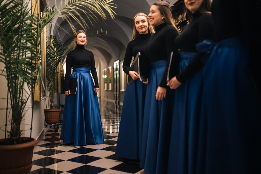 Kobiety w strojach chóralnych stoją w korytarzu z elegancką posadzką i roślinami doniczkowymi. Jedna z nich stoi na pierwszym planie, trzymając nuty, a reszta rozmawia i uśmiecha się