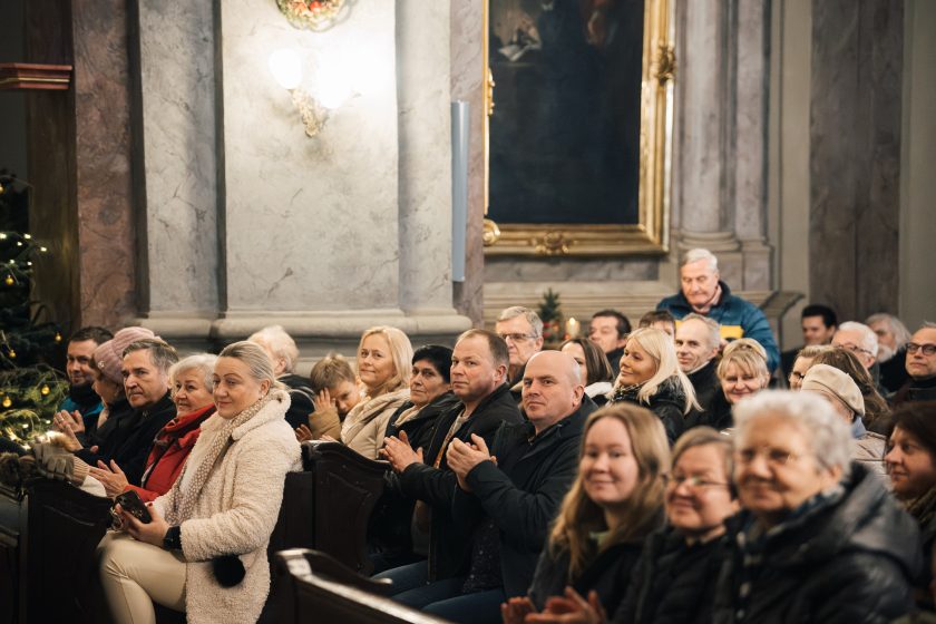 Zgromadzeni widzowie siedzą w ławkach kościoła, oświetleni ciepłym światłem. Na twarzach mają uśmiechy i wyraz zadowolenia, co podkreśla przyjemną atmosferę wydarzenia