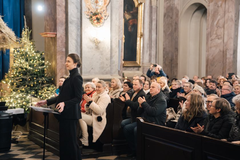 Kobieta w eleganckim czarnym stroju dyryguje chórem, stojąc przed publicznością w kościele. Za nią widać świąteczne ozdoby i chórzystów. Wyraz twarzy wskazuje na pełne zaangażowanie w prowadzenie zespołu