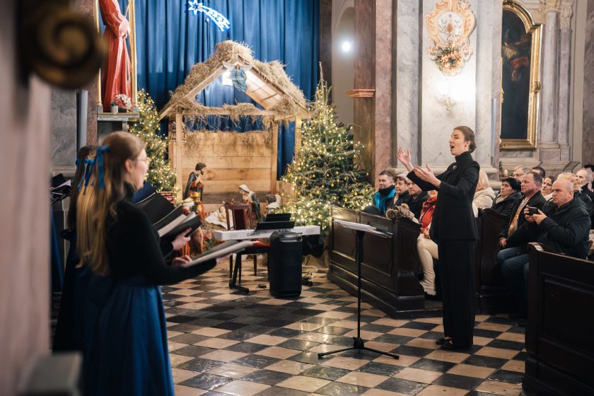 Dyrygentka z uśmiechem i skupieniem stoi na tle pięknie oświetlonej choinki, prowadząc chór podczas koncertu. Jej elegancka postawa podkreśla profesjonalizm i pasję