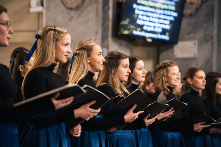 Grupa chórzystek ubranych w czarne bluzki i granatowe spódnice trzyma w rękach nuty. W tle widać wnętrze kościoła z ozdobami świątecznymi i ekranem wyświetlającym tekst. Śpiewają, wyrażając skupienie i emocje