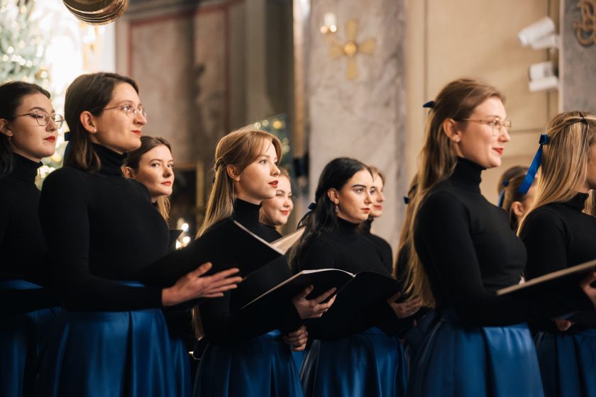 Chórzystki stoją w półprofilu, prezentując zgranie i harmonię. Na twarzach widać profesjonalizm, a otoczenie podkreśla świąteczny nastrój