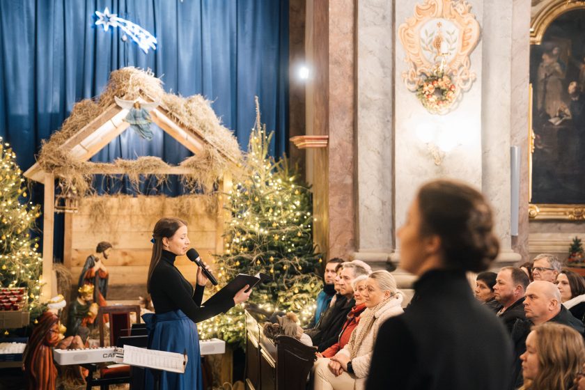 Jedna z chórzystek mówi do mikrofonu, stojąc na tle pięknie udekorowanej szopki. Publiczność siedzi w ławkach, uważnie słuchając. W tle migoczą światełka choinki