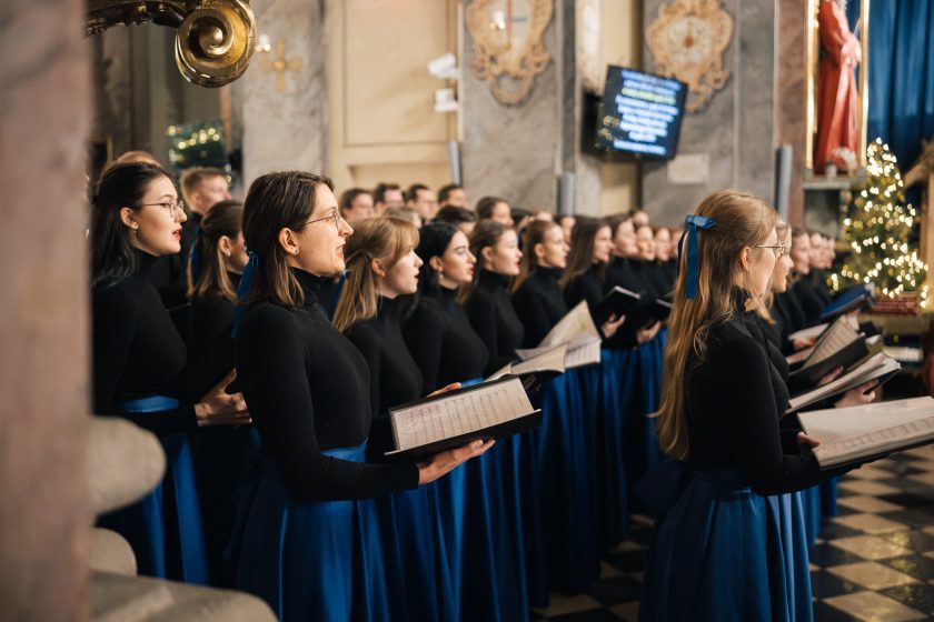 Chórzyści – kobiety i mężczyźni – stoją w rzędach, śpiewając wspólnie. Zgranie i emocje są widoczne w ich postawie i gestach