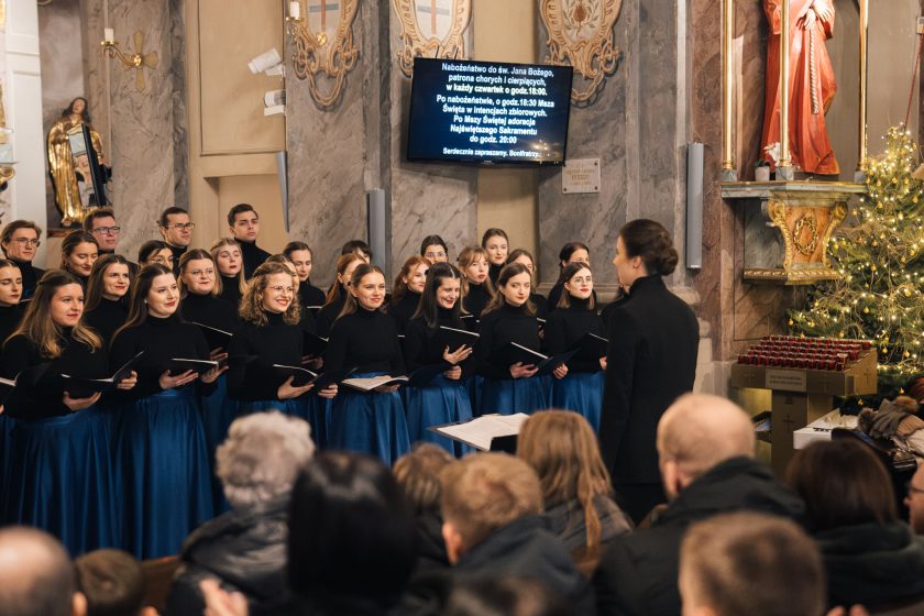 Grupa chórzystów stoi w półokręgu w eleganckich czarnych bluzkach i granatowych spódnicach. Na pierwszym planie widoczna jest dyrygentka w czarnym stroju, prowadząca występ. Tło zdobią świąteczne dekoracje i ekran z informacjami