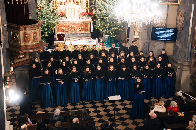 Fragment chóru widziany z lewej strony, chórzystki i chórzyści skupieni na śpiewaniu kolęd. W tle widoczne są świąteczne światełka.