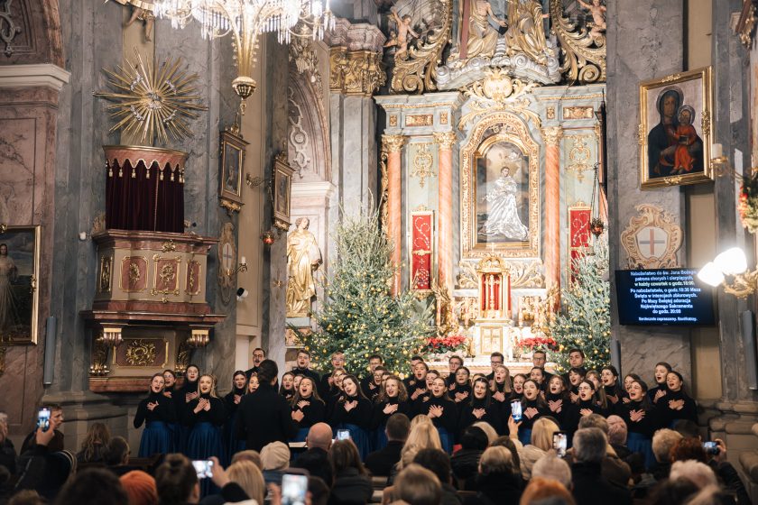 Chór mieszany w eleganckich, czarnych bluzkach i niebieskich spódnicach wykonuje kolędy w barokowym wnętrzu kościoła, ozdobionym świątecznymi dekoracjami i rozświetlonymi choinkami. Na pierwszym planie widoczne są sylwetki publiczności