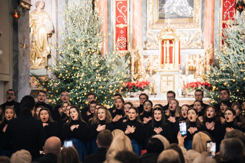 Chór mieszany w eleganckich, czarnych bluzkach i niebieskich spódnicach wykonuje kolędy w barokowym wnętrzu kościoła, ozdobionym świątecznymi dekoracjami i rozświetlonymi choinkami. Na pierwszym planie widoczne są sylwetki publiczności