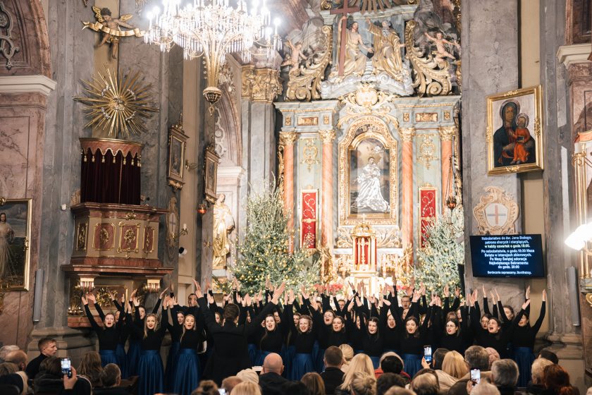 Chór ubrany w ciemne stroje z podniesionymi rękami w geście wyrazu artystycznego. W tle bogato zdobiony ołtarz z dekoracjami bożonarodzeniowymi, w tym dużymi choinkami