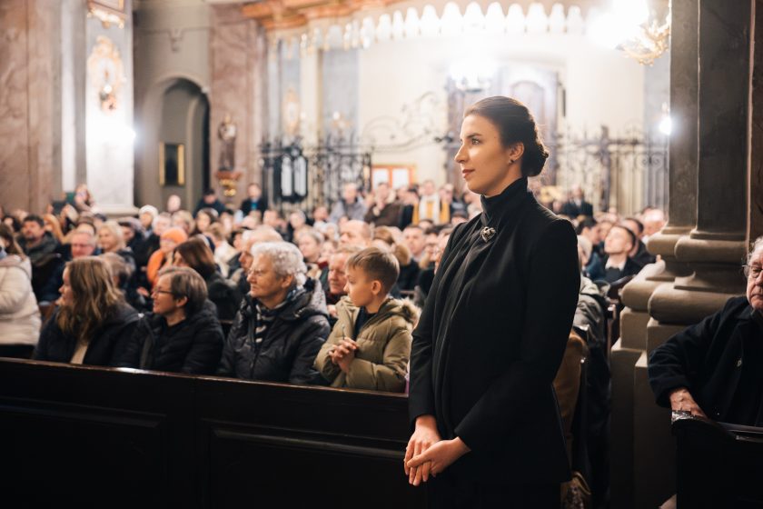 Młoda dyrygentka stoi w ławie kościelnej, w eleganckim czarnym stroju, patrząc spokojnie przed siebie. W tle widoczni są zgromadzeni widzowie wypełniający wnętrze kościoła