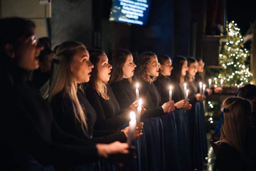 Członkinie chóru śpiewają, trzymając w dłoniach zapalone świece. Nastrojowe światło świec i świąteczne dekoracje tworzą ciepły, intymny klimat