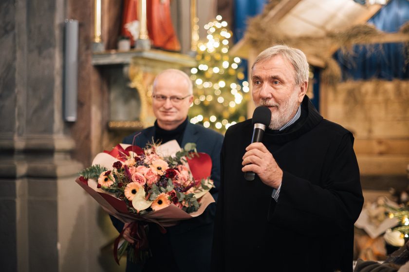 Duchowny ubrany w czarną sutannę przemawia do mikrofonu, stojąc obok mężczyzny trzymającego bukiet kwiatów. W tle dekoracje świąteczne i jasne oświetlenie