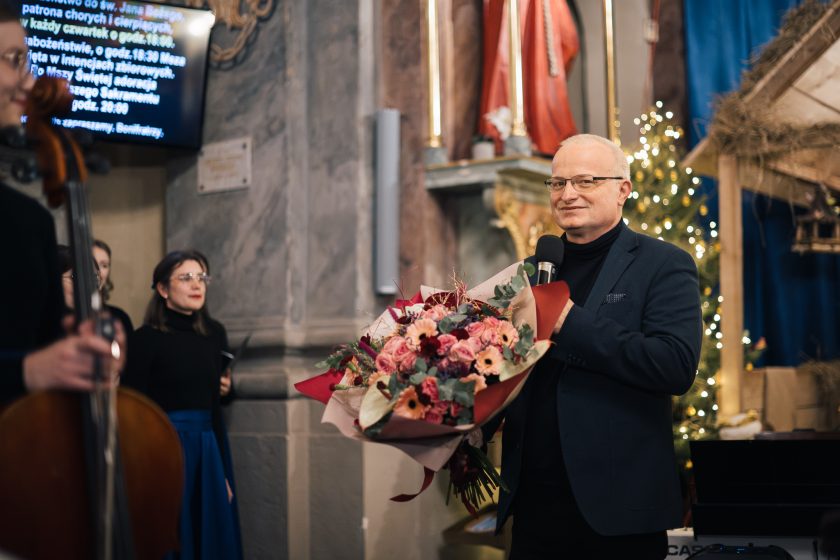 Mężczyzna w ciemnym garniturze trzyma bukiet różowych i czerwonych kwiatów. W tle widoczna jest choinka z ozdobami świątecznymi i fragment ołtarza. Mężczyzna mówi do mikrofonu, uśmiechając się delikatnie