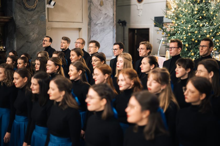 Chór w podniosłym geście, z uniesionymi rękami, występuje przed ołtarzem w bogato zdobionym wnętrzu kościoła, wypełnionym świątecznymi dekoracjami, w tym rozświetlonymi choinkami