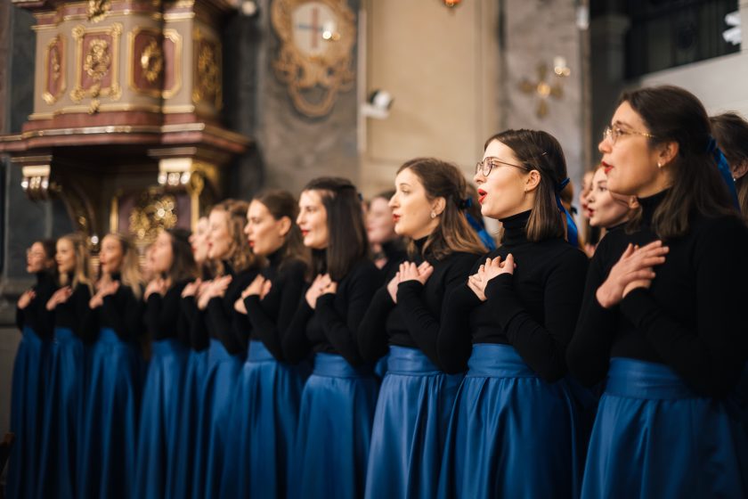 Chór w podniosłym geście, z uniesionymi rękami, występuje przed ołtarzem w bogato zdobionym wnętrzu kościoła, wypełnionym świątecznymi dekoracjami, w tym rozświetlonymi choinkami. Widownia siedzi na ławkach