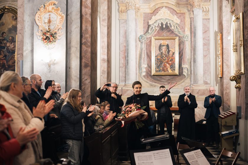 Kobieta w eleganckim garniturze trzymająca bukiet kwiatów, przemawia do mikrofonu na tle oświetlonej choinki i dekoracji świątecznych w kościele. W tle widać inne osoby oraz drewnianą szopkę.