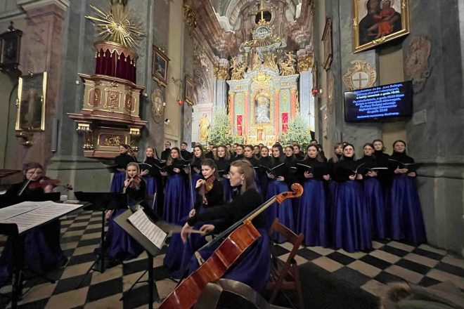 Dyrygentka prowadząca chór podczas koncertu. Na pierwszym planie muzycy orkiestry smyczkowej, a w tle chórzyści oraz oświetlony ołtarz z bożonarodzeniowymi dekoracjami