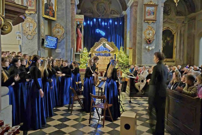 Chórzystki schodzące ze sceny po zakończeniu występu. W tle widać szopkę bożonarodzeniową, udekorowane choinki oraz zgromadzoną publiczność, która gromkimi brawami wyraża swoje uznanie