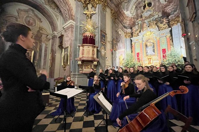 Chór wykonujący utwory przed majestatycznym ołtarzem, w towarzystwie orkiestry smyczkowej. W tle rozświetlone dekoracje bożonarodzeniowe oraz barokowa architektura kościoła