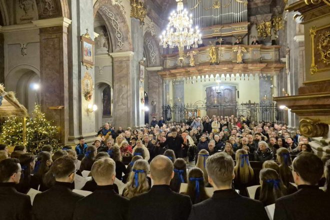 Widok na pełną publiczność zgromadzoną w pięknie zdobionym wnętrzu kościoła podczas występu chóru. W tle widać oświetlone organy, barokowe zdobienia i żyrandol nadający uroczystego charakteru wydarzeniu