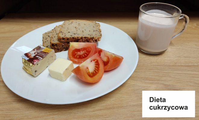 Na stole znajduje się talerz z elementami diety cukrzycowej (napis na kartce: „Dieta cukrzycowa”). Na talerzu podano dwie kromki chleba pełnoziarnistego, kawałek sera żółtego, porcję twarożku kremowego, oraz świeże pomidory pokrojone w ćwiartki. Obok znajduje się przezroczysty kubek z mlekiem