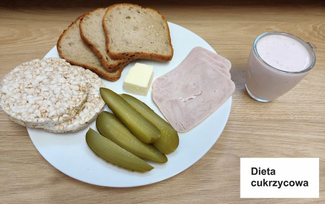 Zdjęcie przedstawia kolację w diecie cukrzycowej, oznaczonej na białej karteczce z napisem "Dieta cukrzycowa". Na talerzu znajdują się trzy kromki chleba razowego, ryżowe wafle, kawałek masła, plasterki wędliny drobiowej oraz kilka kawałków ogórków konserwowych. Obok talerza stoi szklanka jogurtu lub koktajlu owocowego