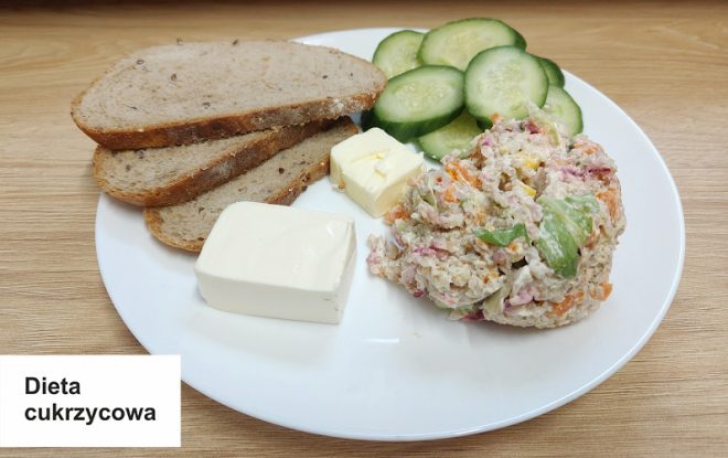 Kolacja w diecie cukrzycowej, oznaczonej na białej karteczce z napisem "Dieta cukrzycowa". Na talerzu znajdują się trzy kromki chleba razowego, kawałek masła, porcja twarogu, kilka plasterków świeżego ogórka oraz sałatka jarzynowa z majonezem i warzywami. Wszystko ułożone estetycznie na białym talerzu