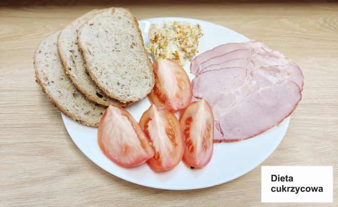 Talerz przedstawia posiłek w postaci trzech kromek chleba żytniego, plasterków pomidora, wędliny oraz pasty jajecznej. Obok na stole umieszczona jest kartka z napisem „Dieta cukrzycowa”