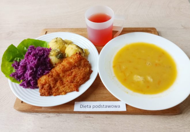 Zdjęcie przedstawia posiłek z diety podstawowej, ułożony na drewnianej tacy. Na białym talerzu znajduje się panierowany filet drobiowy, purée ziemniaczane posypane koperkiem oraz surówka z czerwonej kapusty podanej na liściu sałaty. Obok widać głęboki talerz z żółtą zupą jarzynową. Do posiłku dołączono kubek z różowym napojem