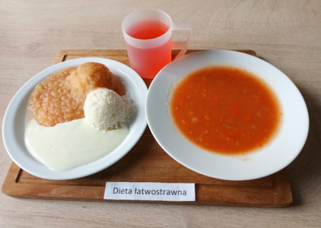 Na drewnianej desce znajduje się łatwostrawny obiad w porcelanowych talerzach (napis na kartce: „Dieta łatwostrawna”). Na jednym talerzu podano ryż w formie kulki, pulpeta z delikatnym sosem i mus jabłkowy. Na drugim talerzu znajduje się klarowna zupa pomidorowa z kawałkami warzyw. Całość uzupełnia różowy napój w plastikowym kubku