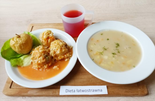 Na drewnianej tacce widoczny jest zestaw obiadowy przygotowany w ramach diety łatwostrawnej, co podkreśla umieszczona poniżej kartka z napisem „Dieta łatwostrawna”. Na białym talerzu znajdują się trzy gołąbki ryżowe w delikatnym sosie pomidorowym oraz pieczone jabłko na liściu sałaty. Obok stoi miska z jasną zupą jarzynową, posypaną drobno posiekanym koperkiem. Całość uzupełnia plastikowy kubek z czerwonym napojem