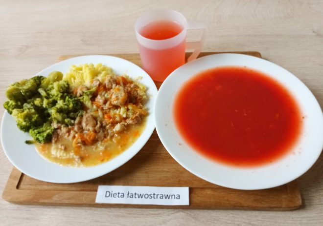 Na drewnianej tacy ustawiono zestaw obiadowy oznaczony jako "Dieta łatwostrawna". Na białym talerzu znajduje się gotowany brokuł, ziemniaczane puree oraz delikatny gulasz z mięsa mielonego i drobno pokrojonej marchewki w sosie. Obok podano głęboki talerz z czerwoną zupą pomidorową, a całość dopełnia przezroczysty kubek z różowym kompotem